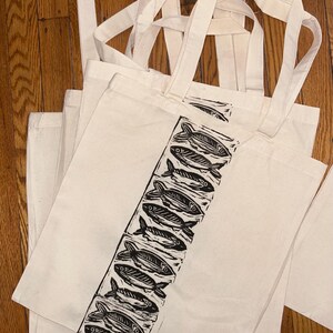 May include: Several off-white canvas tote bags with long handles are stacked on a wooden surface. Each bag features a vertical black and white fish print design. The bags are ideal for carrying groceries or other items.