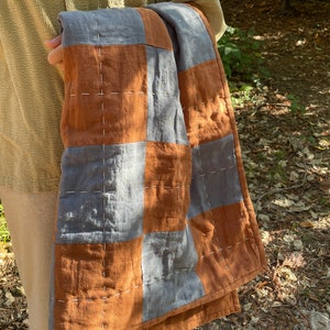 May include: A brown and gray patchwork quilt with white stitching. The quilt is folded and held by a person.