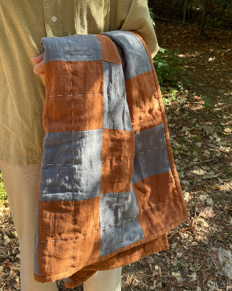 May include: A brown and gray patchwork quilt with white stitching. The quilt is folded and held by a person.