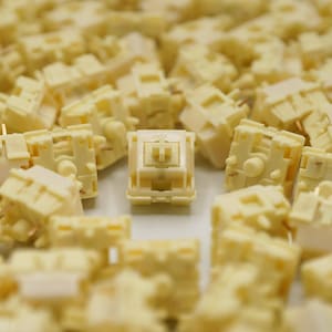May include: A close-up of a group of cream-colored mechanical keyboard keycaps. One keycap is in focus and has a cross-shaped stem.