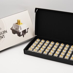 以下が含まれることがあります： 黒色の箱の中に、白と金のメカニカルキーボードスイッチが入っています。箱が開いていて、スイッチが見えています。箱には「Akko Penguin Silent」とラベルが貼られています。