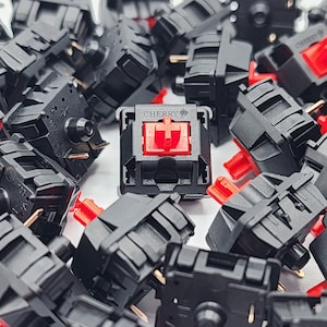 May include: A close-up of a group of black mechanical keyboard keycaps with red stems. The keycaps are arranged in a random pattern, and one keycap is labelled "CHERRY".