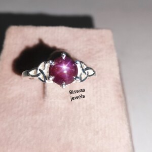 Peut inclure: Une bague en argent avec une pierre précieuse en rubis étoilé rouge. La bague est ornée d'un motif de nœud celtique. Le texte "Biswas jewels" est visible sous la bague.