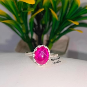 May include: A silver ring with a pink star ruby gemstone. The ring has a decorative border of small white stones. The text "Biswas Jewels" is visible in the image.