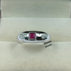 May include: A silver ring featuring a red square-shaped gemstone and two smaller white gemstones. The ring is displayed in a white box with the inscription "Biswas Jewels" at the bottom.