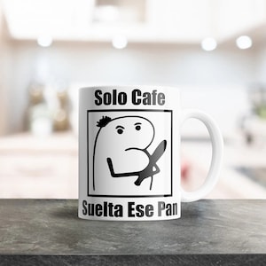Pode incluir: Caneca de cerâmica branca com uma ilustração em preto e branco de uma pessoa com uma expressão de raiva. O texto "Solo Cafe" e "Suelta Ese Pan" está impresso na caneca.