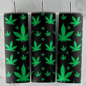 May include: Three black insulated tumblers with a repeating pattern of bright green cannabis leaves. Each tumbler has a silver-colored straw and a black lid. The background is a light marble pattern.