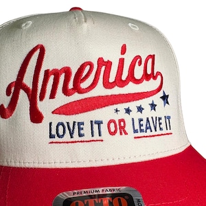 May include: A baseball cap with a white crown and a red brim. The word "America" is embroidered in red script, with "LOVE IT OR LEAVE IT" in blue block letters. The design includes blue stars.