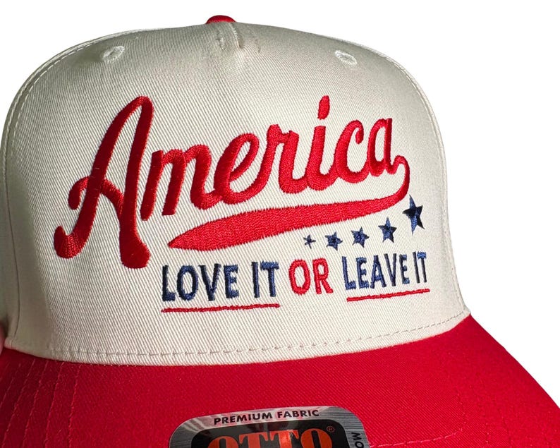May include: A baseball cap with a white crown and a red brim. The word "America" is embroidered in red script, with "LOVE IT OR LEAVE IT" in blue block letters. The design includes blue stars.