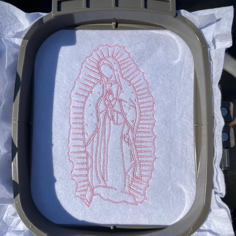 May include: Embroidered image of the Virgin Mary in a pink outline on a white fabric. The image is in a hoop for embroidery.