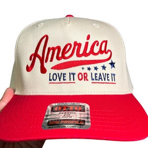 May include: A white and red baseball cap with the word "America" embroidered in red script. The text "LOVE IT OR LEAVE IT" is below the word "America". The cap has a red brim and a tag that reads "CLASSIC FIT".