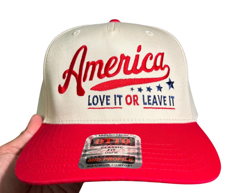 May include: A white and red baseball cap with the word "America" embroidered in red script. The text "LOVE IT OR LEAVE IT" is below the word "America". The cap has a red brim and a tag that reads "CLASSIC FIT".