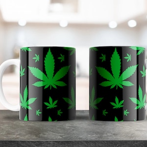 May include: Two white ceramic mugs with black and green marijuana leaf designs. The mugs are on a gray countertop.