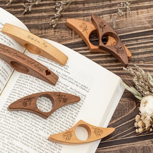 May include: Several wooden book page holders are displayed on an open book. The holders are in various shades of brown and tan, some with floral designs and text like "With love, Joy" and "Sisters forever and always".