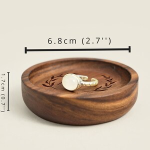 May include: A wooden ring dish with a leafy design engraved on the surface. The dish is 6.8 inches in diameter and 0.7 inches tall. A silver ring with a white stone is resting on the dish.