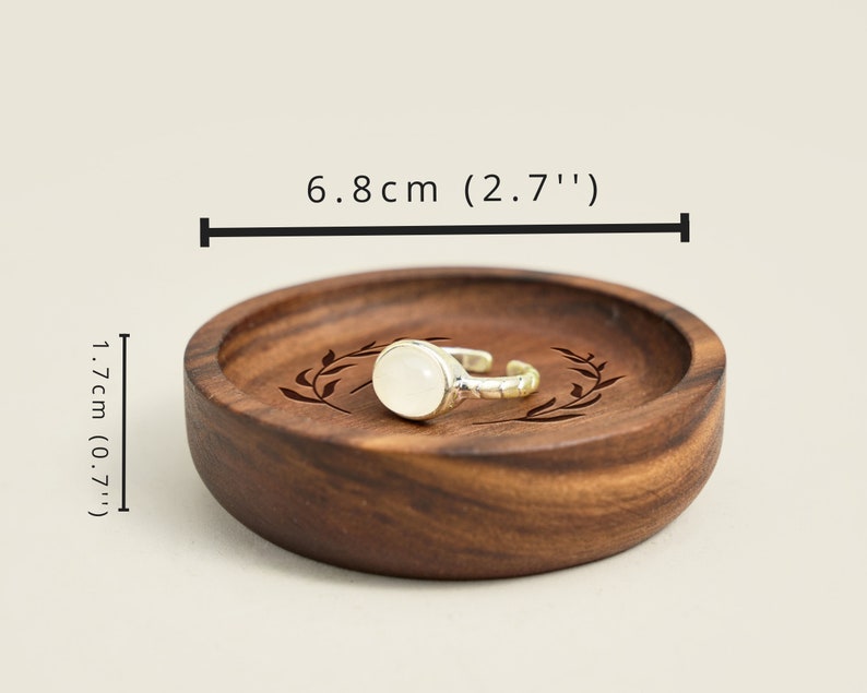 May include: A wooden ring dish with a leafy design engraved on the surface. The dish is 6.8 inches in diameter and 0.7 inches tall. A silver ring with a white stone is resting on the dish.