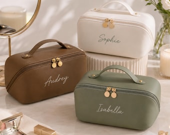 Puede incluir: Tres neceseres rectangulares con asas superiores y cierres de doble cremallera. Los neceseres son de color blanco, marrón y verde salvia, cada uno con un nombre personalizado en letra cursiva: Sophie, Audrey e Isabella.