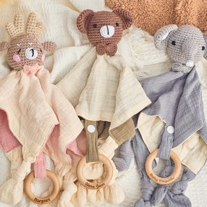 K&ouml;nnte beinhalten: Drei geh&auml;kelte Baby-Sicherheitsdecken mit Tierkopf-Anh&auml;ngseln. Die Decken sind beige mit rosa, gr&uuml;nem oder grauem Besatz. Jede Decke hat einen Holzring mit einem eingravierten Namen. Die Namen sind Margaux, Nora Jane und Baby Kojyunki.