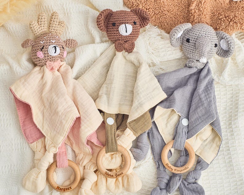 K&ouml;nnte beinhalten: Drei geh&auml;kelte Baby-Sicherheitsdecken mit Tierkopf-Anh&auml;ngseln. Die Decken sind beige mit rosa, gr&uuml;nem oder grauem Besatz. Jede Decke hat einen Holzring mit einem eingravierten Namen. Die Namen sind Margaux, Nora Jane und Baby Kojyunki.