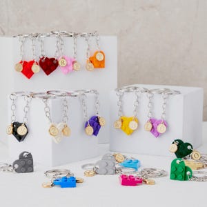 Può includere: Una collezione di portachiavi a forma di cuore in vari colori, tra cui rosso, rosa e giallo. Ogni portachiavi presenta un ciondolo con iniziale dorata e una catena argentata. Alcuni portachiavi sono fatti di mattoncini ad incastro.