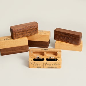 May include: Wooden ring boxes in various styles. Some are engraved with names and dates. One box is open, displaying two rings. The text on the open box reads "The Solomon Begins" and "Barry & Kevin November 12, 2023".