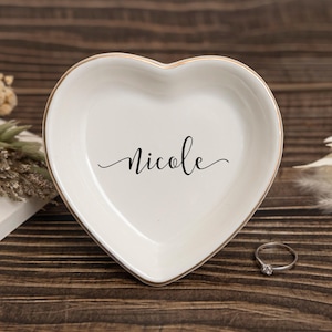 May include: A heart-shaped, white ceramic trinket dish with a gold rim and the name "Nicole" in black script. A silver ring with a clear stone sits nearby on a wooden surface. Decorative dried flowers are in the background.