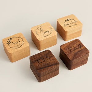 Könnte beinhalten: Fünf Holzkisten für Ringe mit gravierten Namen und Daten. Die Kisten sind aus hellem und dunklem Holz.