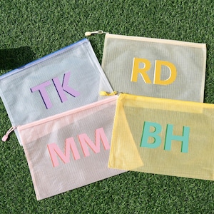 May include: Four mesh zipper pouches in pastel shades: light blue, pink, yellow, and beige. Each pouch has large, block letter initials in contrasting colours: purple, pink, yellow, and teal. The pouches are arranged on a green surface.