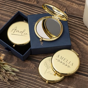 May include: Three round gold-tone compact mirrors with hinged lids. One mirror is open, revealing a reflective surface. Two closed mirrors feature black engraved text reading Amelia and floral line art designs. They are presented in navy blue boxes.