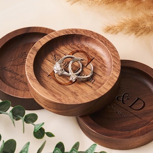 May include: Three wooden ring dishes with engraved initials and dates. The dishes are made of dark wood and have a smooth, polished finish.