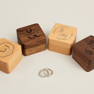 May include: Four wooden ring boxes with engraved designs and a pair of silver rings. The boxes are square-shaped, with two made of light wood and two of dark wood. The boxes have personalized text and floral designs.