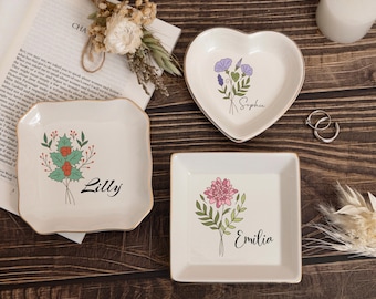 May include: Three decorative ceramic trinket dishes with gold trim. One is heart-shaped, one square, and one rectangular. Each dish features a floral design and a name: Lilly, Sophia, and Emilia. The dishes are on a wooden surface.