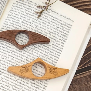 May include: Two wooden book page holders, one dark brown and one light brown, rest on an open book. Each has a circular opening and floral engraving. The book is open to a page of text.