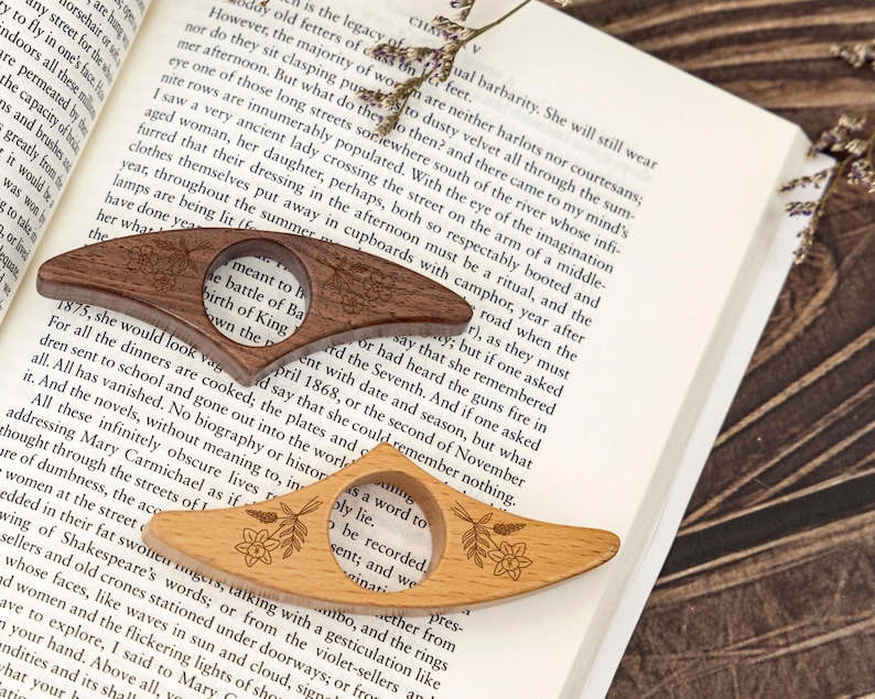 May include: Two wooden book page holders, one dark brown and one light brown, rest on an open book. Each has a circular opening and floral engraving. The book is open to a page of text.