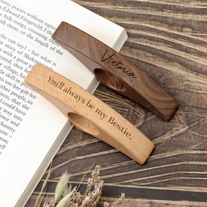 May include: Two wooden book page holders. One is light-colored with the text "You'll always be my Bestie." The other is dark-colored with the name "Victoria." Both have a curved design to hold pages open. They rest on a book.