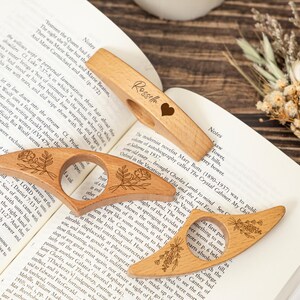 May include: Three wooden thumb page holders with floral designs and a heart. One has the name "Rossella" engraved. These book accessories are designed to keep pages open while reading. Perfect for book lovers.
