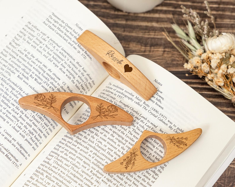 May include: Three wooden thumb page holders with floral designs and a heart. One has the name "Rossella" engraved. These book accessories are designed to keep pages open while reading. Perfect for book lovers.
