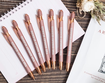 May include: Six rose gold pens with the text "THE SUNDAY SCARIES" printed on the barrel. The pens are arranged on a spiral notebook. The pens have gold-colored accents and are on a wooden surface.