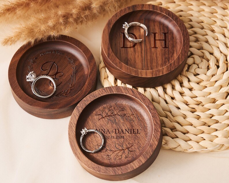 May include: Three wooden ring dishes with engraved designs. The first dish has the initials "D & H" engraved on it. The second dish has the initials "D" and a floral design engraved on it. The third dish has the text "Anna + Daniel 12.12.2023" and a floral design engraved on it.