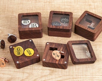 May include: A collection of personalized wooden boxes and cufflinks. The boxes are square with clear tops, displaying engraved initials and names. Cufflinks are gold and silver-toned, also engraved with initials. The items are arranged on a light wood surface.