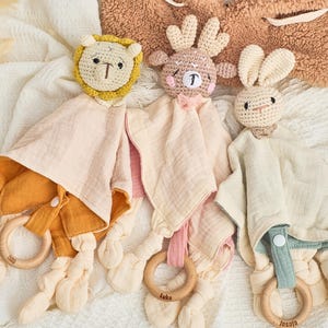 K&ouml;nnte beinhalten: Drei geh&auml;kelte Babyspielzeuge mit befestigten Decken. Die Spielzeuge sind ein L&ouml;we, ein Rentier und ein Kaninchen. Jedes Spielzeug hat einen Holzring, der an der Decke befestigt ist.