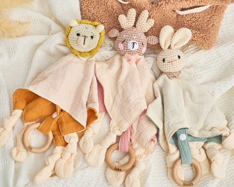 K&ouml;nnte beinhalten: Drei geh&auml;kelte Babyspielzeuge mit befestigten Decken. Die Spielzeuge sind ein L&ouml;we, ein Rentier und ein Kaninchen. Jedes Spielzeug hat einen Holzring, der an der Decke befestigt ist.