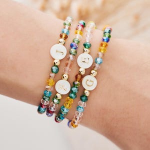 May include: Four colourful beaded bracelets with gold accents and white letter charms. The bracelets feature different coloured beads, including pink, blue, green, and yellow.