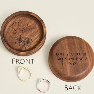 May include: A wooden ring box with a personalized inscription on the lid. The front of the box is engraved with the name "Sophia" and a four-leaf clover design. The back of the box is engraved with the message "Love you to the moon and back! -Naz".