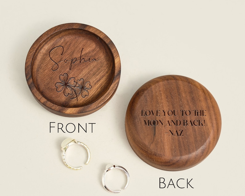 May include: A wooden ring box with a personalized inscription on the lid. The front of the box is engraved with the name "Sophia" and a four-leaf clover design. The back of the box is engraved with the message "Love you to the moon and back! -Naz".