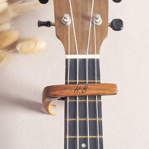 Pode incluir: Um capo de madeira com as iniciais "H.B." gravadas nele, preso ao braço de um ukulele.