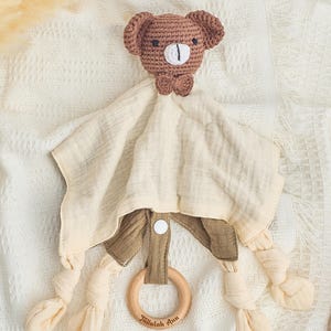 K&ouml;nnte beinhalten: Ein brauner geh&auml;kelter B&auml;r mit einer Schleife ist an einer beigefarbigen Musselin-Babydecke mit einem Holz-Bei&szlig;ring befestigt. Der Ring tr&auml;gt den Text "Tallaulah Ann" eingraviert.