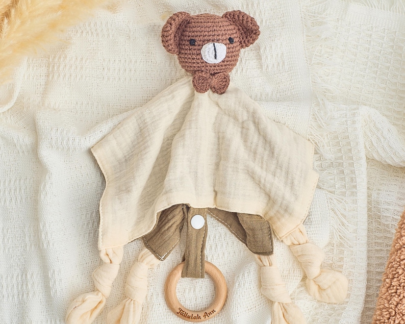 K&ouml;nnte beinhalten: Ein brauner geh&auml;kelter B&auml;r mit einer Schleife ist an einer beigefarbigen Musselin-Babydecke mit einem Holz-Bei&szlig;ring befestigt. Der Ring tr&auml;gt den Text "Tallaulah Ann" eingraviert.