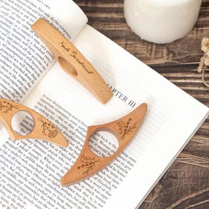 May include: Three wooden thumb page holders with floral engravings and a hole for the thumb rest on an open book. One holder has the name "Kade Barnbrook" engraved. The wood has a light brown color.