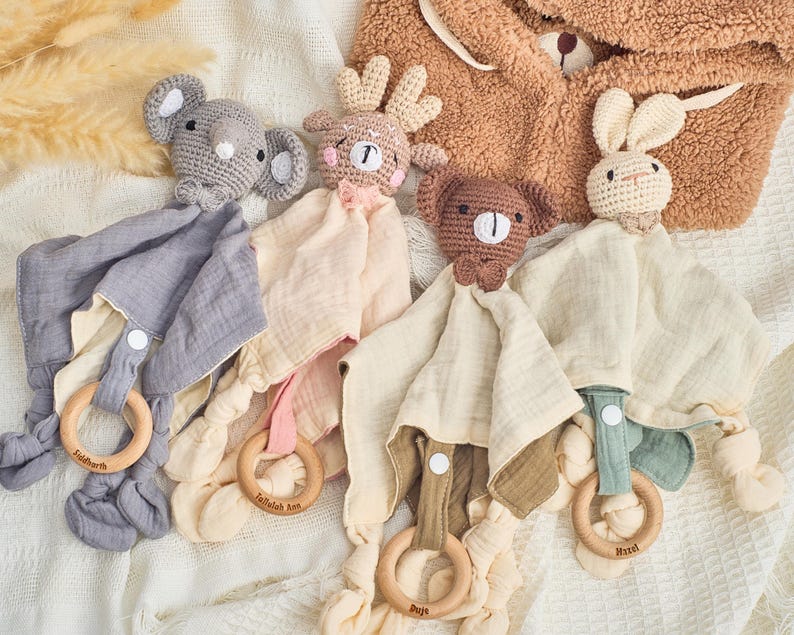 K&ouml;nnte beinhalten: Vier geh&auml;kelte Tier-Babyspielzeuge mit befestigten Musselin-T&uuml;chern. Die Spielzeuge sind ein grauer Elefant, ein braunes Reh, ein brauner Hund und ein wei&szlig;es Kaninchen. Jedes Spielzeug hat einen Holzring, der am Tuch befestigt ist.